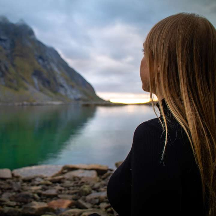 femeie în jachetă neagră așezată pe stâncă lângă lac puzzle online