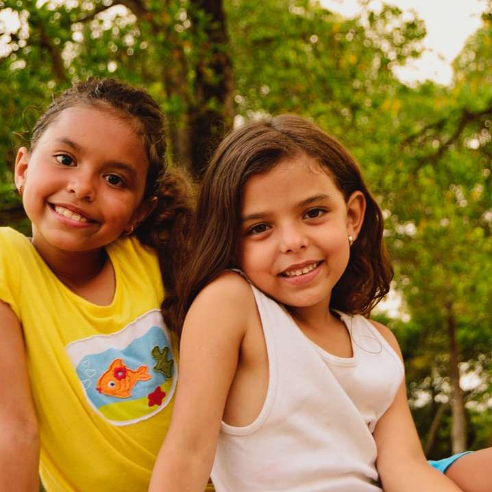 2 niñas sonriendo y de pie cerca de árboles verdes durante el día puzzle deslizante online