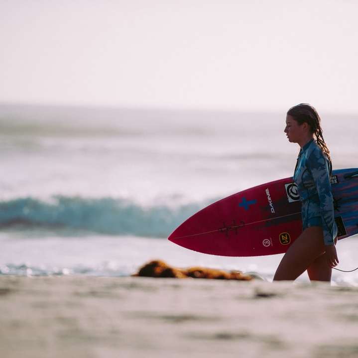 Frau, die rotes und blaues Surfbrett trägt, das am Ufer geht Schiebepuzzle online