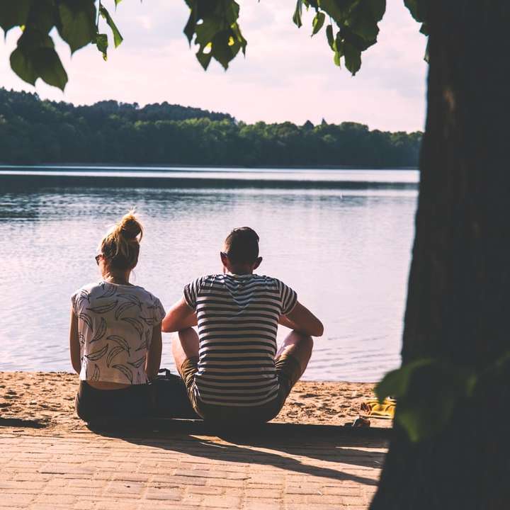 men and woman sitting on pavement facing body of water sliding puzzle online