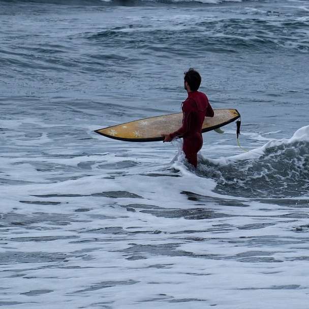 man i röd och svart våtdräkt som håller den vita surfbrädan Pussel online
