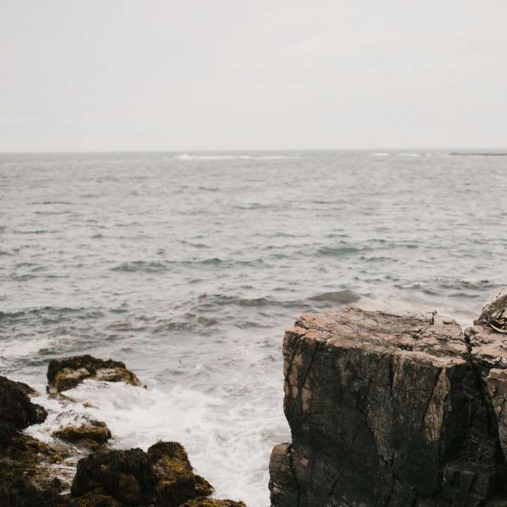 Mann im blauen Hemd, das auf Felsformation nahe Meer sitzt Schiebepuzzle online
