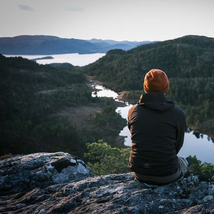 uomo in felpa con cappuccio nera seduto sulla roccia guardando il lago puzzle online