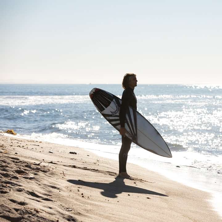 man i svart våt kostym som håller den vita surfbrädan Pussel online