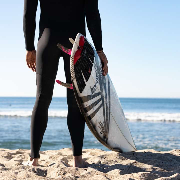 Persona con tabla de surf blanca y azul de pie en la playa puzzle deslizante online
