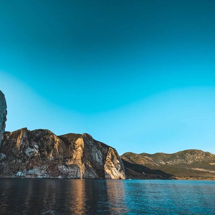 barna sziklás hegy kék tenger mellett, kék ég alatt csúszó puzzle online