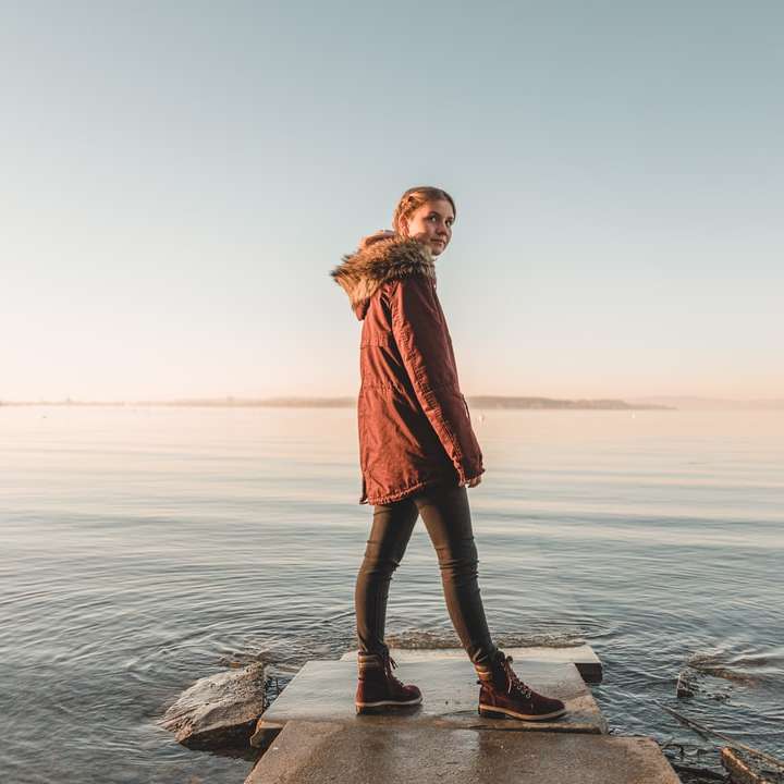 жінка в червоному пальті стоїть на скелі біля моря вдень онлайн пазл