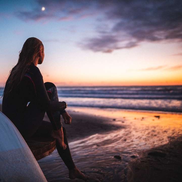 kobieta w czarnej kurtce siedzi na białej desce surfingowej na plaży puzzle przesuwne online