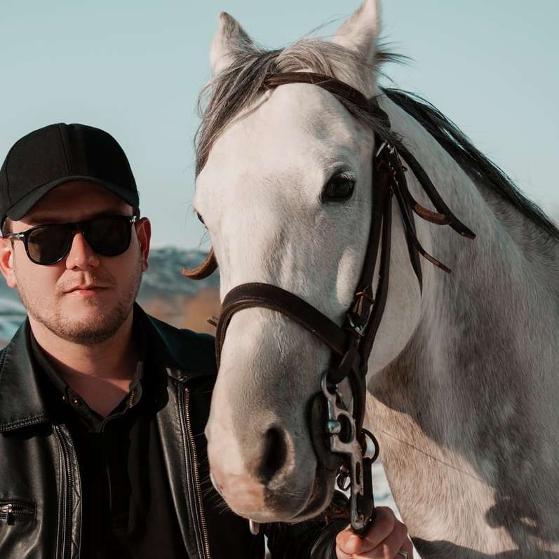 Hombre con chaqueta de cuero negro de pie junto a caballo blanco rompecabezas en línea