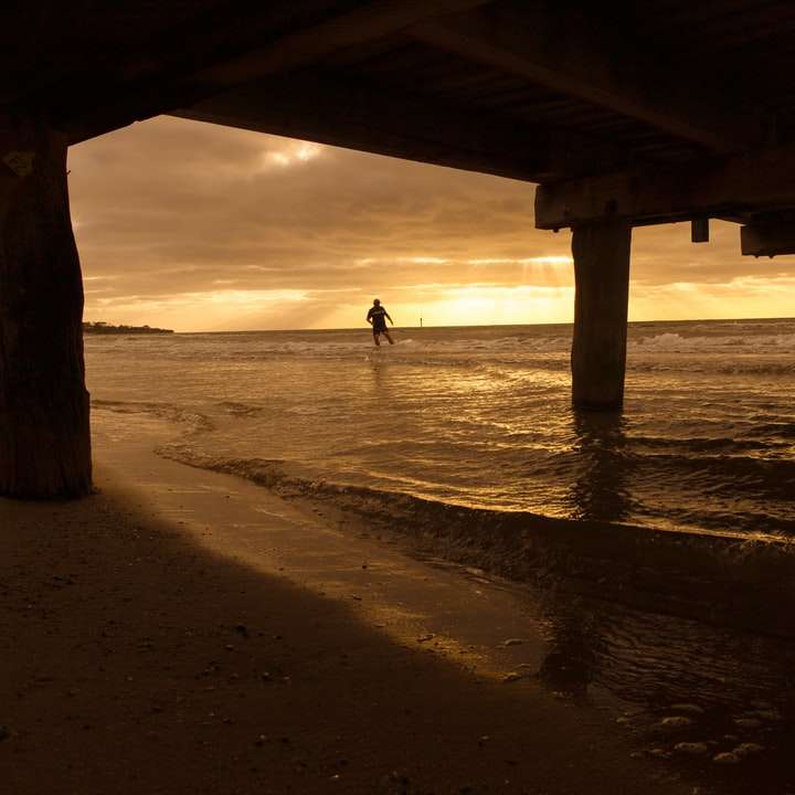 Persona caminando en la playa bajo el puente de madera durante el día rompecabezas en línea