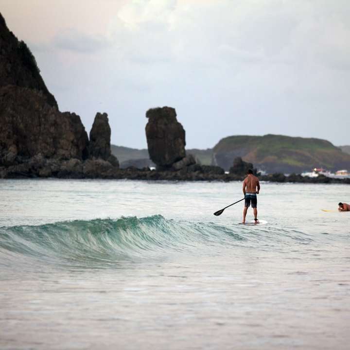 mężczyzna w czarnych spodenkach stojący na białej desce surfingowej na morzu puzzle przesuwne online