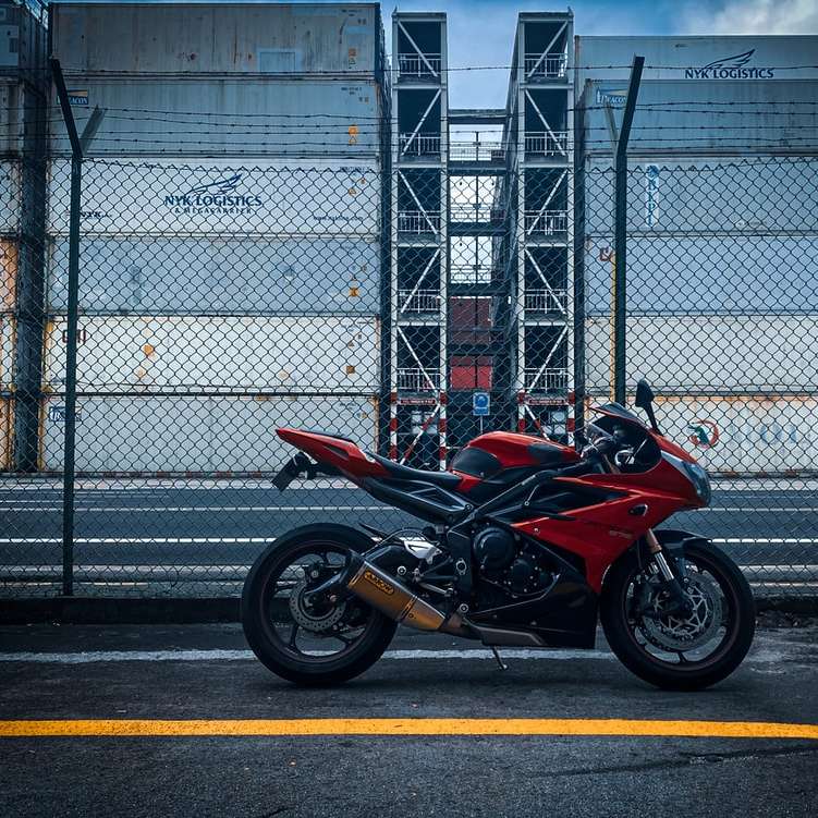 красно-черный спортивный мотоцикл припаркован у серого металлического забора онлайн-пазл