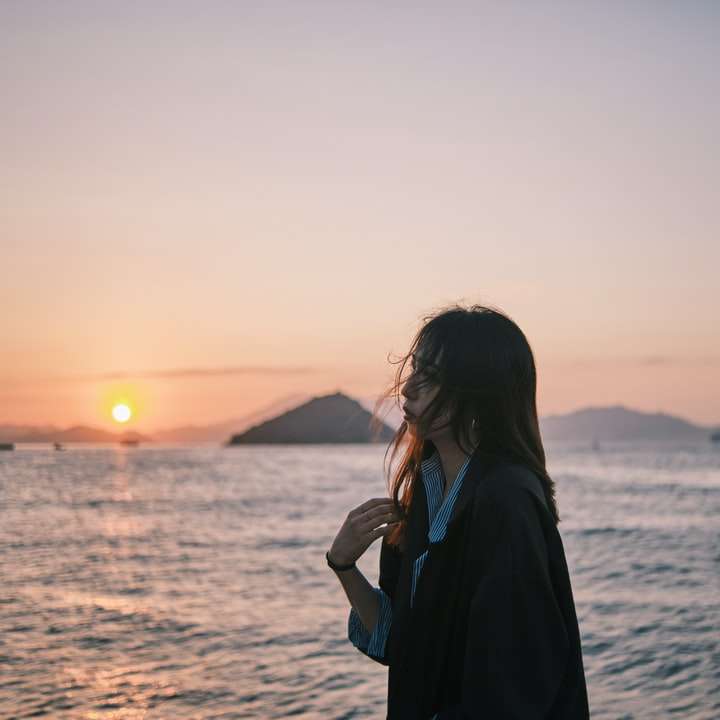 vrouw in zwarte jas staande op het strand tijdens zonsondergang schuifpuzzel online