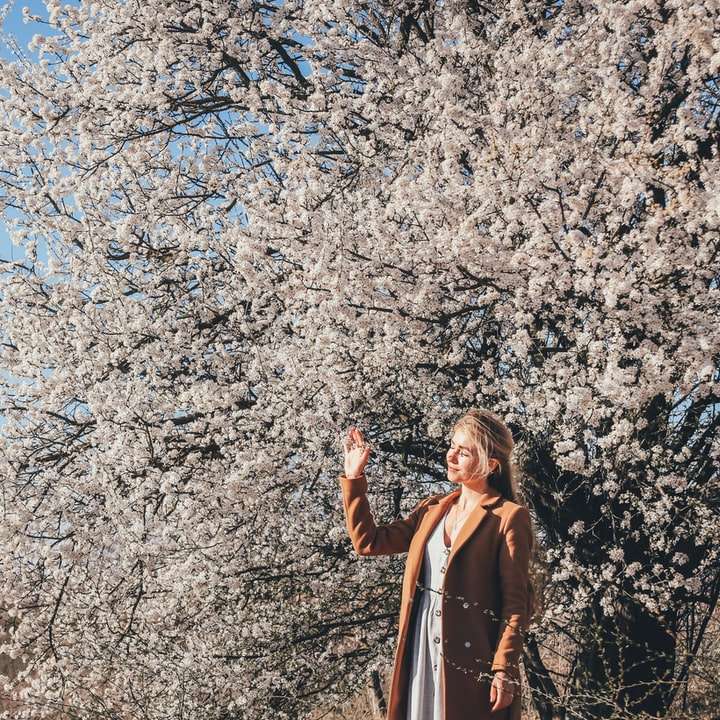 woman in brown coat standing near white cherry blossom tree sliding puzzle online