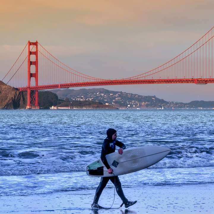 Silueta del hombre que sostiene la tabla de surf en la playa durante la puesta del sol rompecabezas en línea