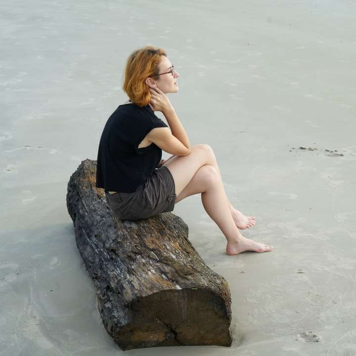 Γυναίκα σε μαύρη δεξαμενή κορυφή κάθεται σε γκρίζο βράχο δίπλα στη θάλασσα συρόμενο παζλ online