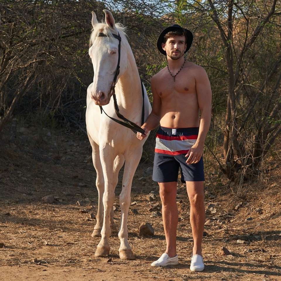Homme topless en short bleu debout à côté de cheval blanc puzzle en ligne