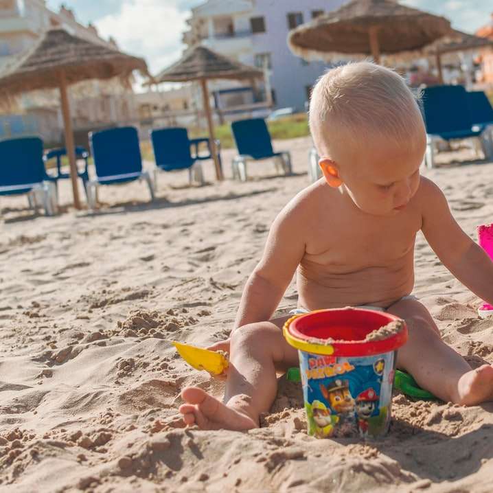 Menino de topless segurando balde de plástico azul na areia puzzle deslizante online