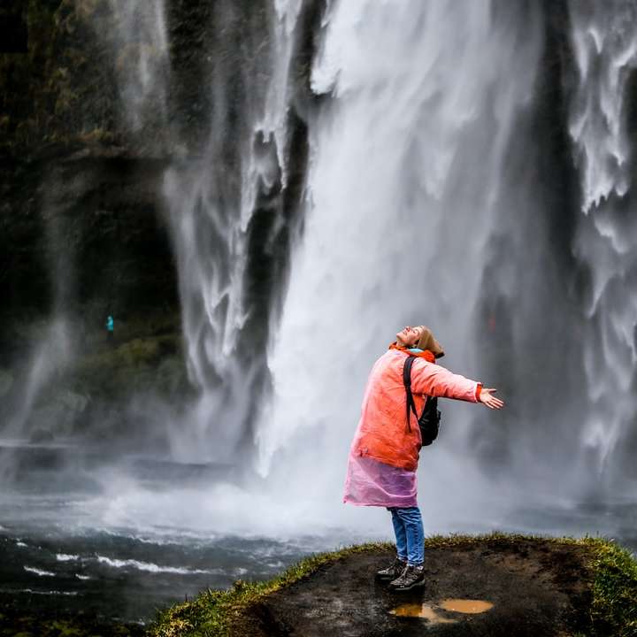 Frau in blauer Jacke, die auf Felsen in der Nähe von Wasserfällen steht Online-Puzzle