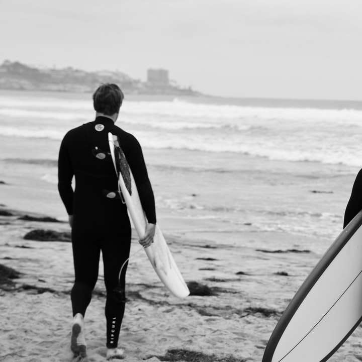 mężczyzna w czarnej kurtce trzymający białą deskę surfingową spacerujący po plaży puzzle przesuwne online