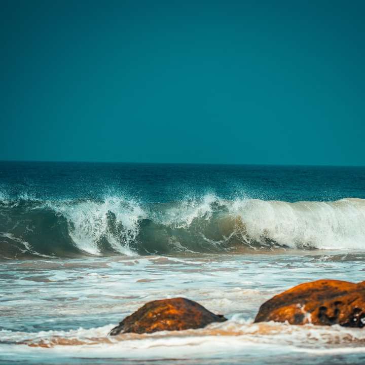 havsvågor som slår mot brun klippformation under blå himmel Pussel online