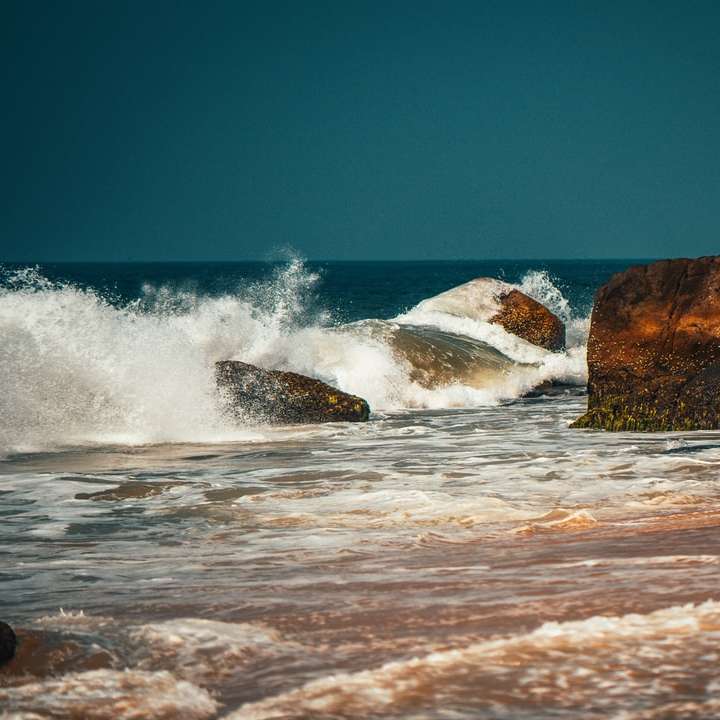океанські хвилі розбиваються на коричневе скельне утворення під блакитним небом розсувний пазл онлайн