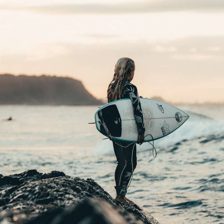 kobieta w czarnym kombinezonie nosząca białą deskę surfingową puzzle online
