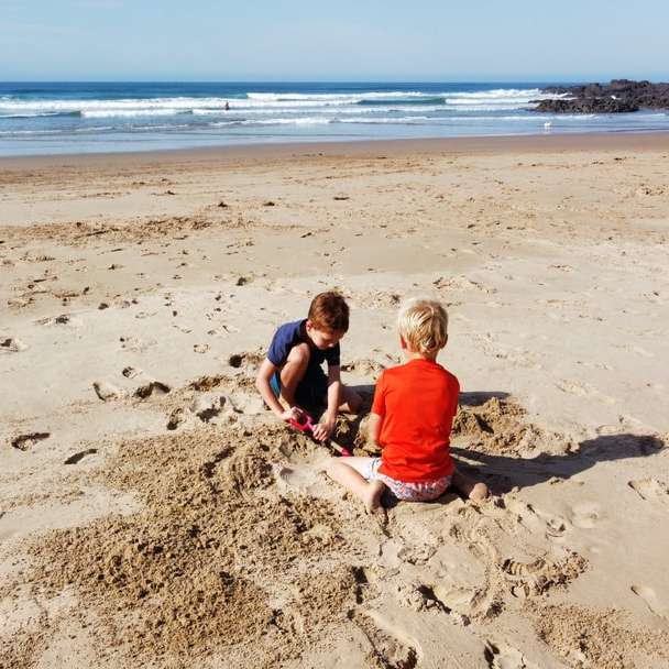 2 chłopców siedzących na brązowym piasku w pobliżu morza w ciągu dnia puzzle online
