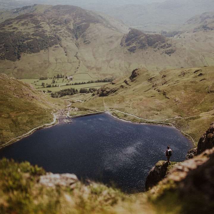 човек в черно яке, стоящ на скално образувание близо до езерото онлайн пъзел